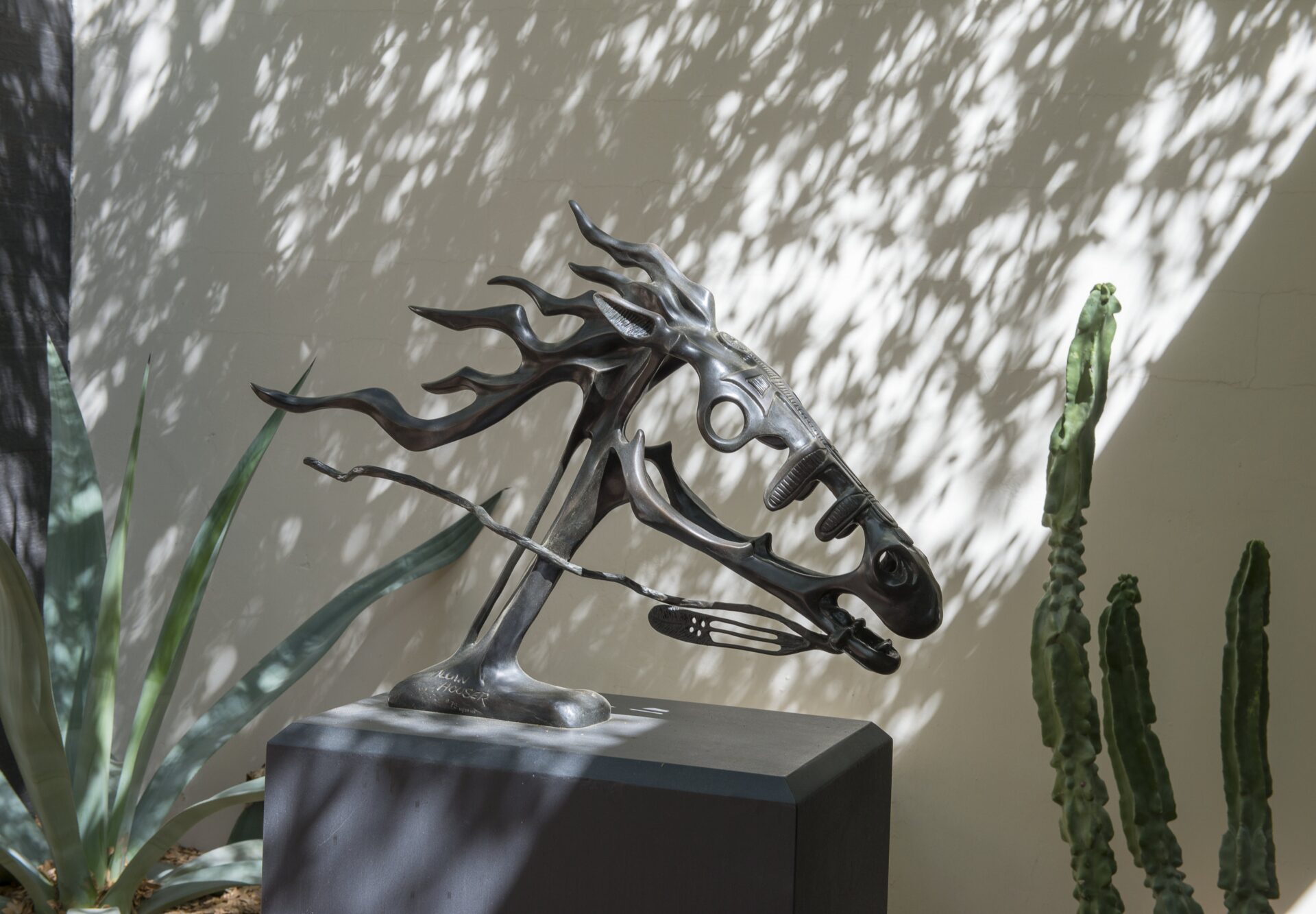 Metal horse head sculpture with flowing mane displayed on a pedestal, surrounded by leafy plants and dappled sunlight.