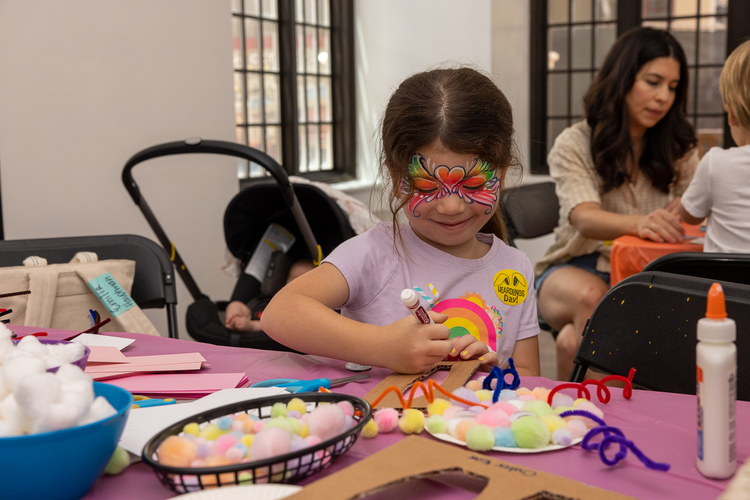 A child with face paint is crafting with colorful pom-poms and pipe cleaners. An adult and a baby are in the background.