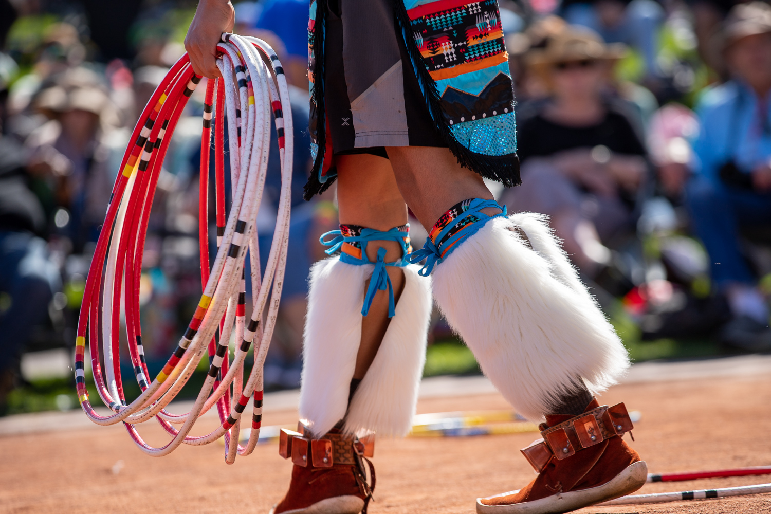 A person holding colorful hoops is dressed in traditional attire with fur leggings and beaded decorations, performing at an outdoor event.