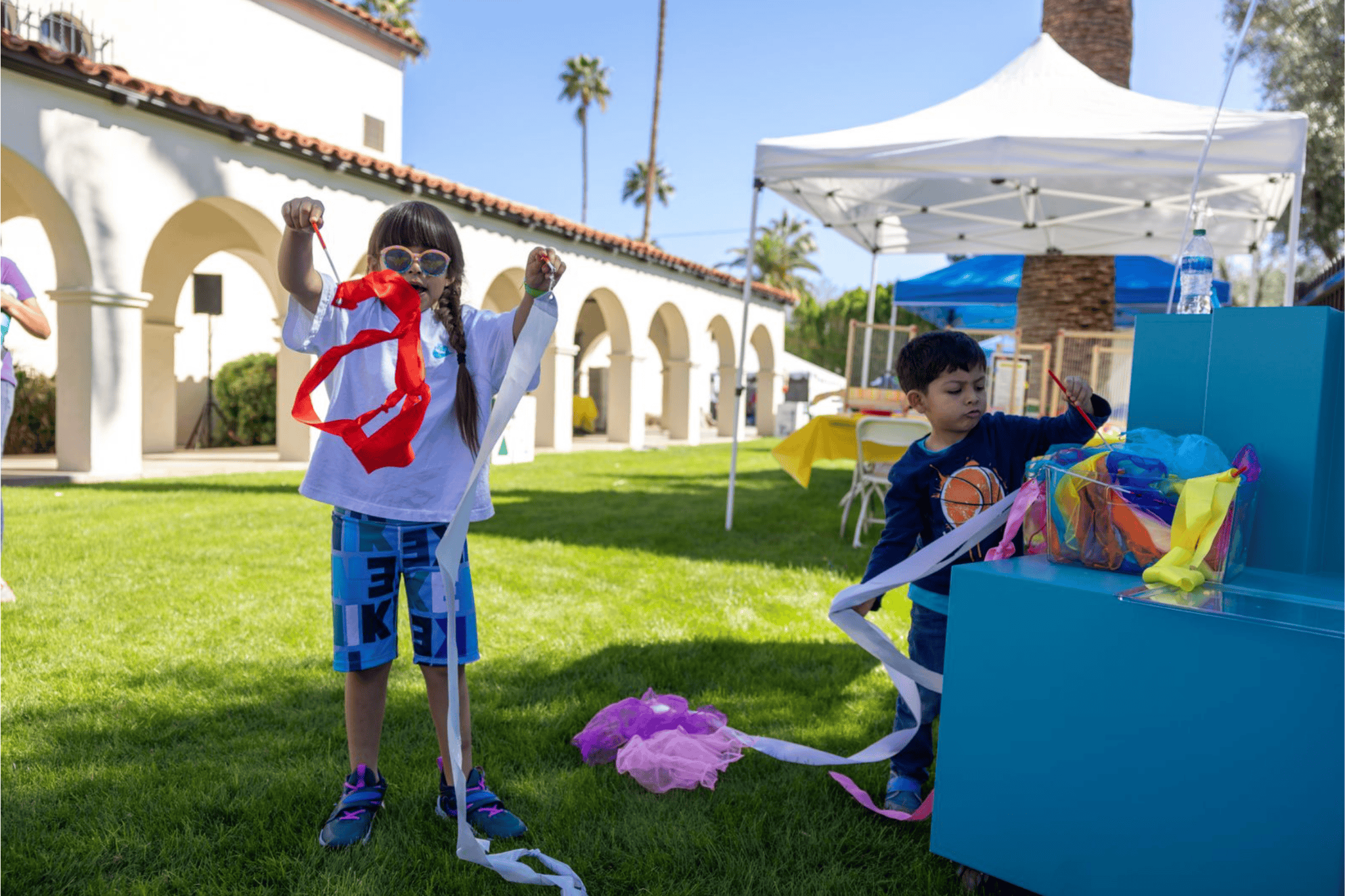 Two children play with colorful streamers outdoors near a booth and a building with arches on a sunny day.