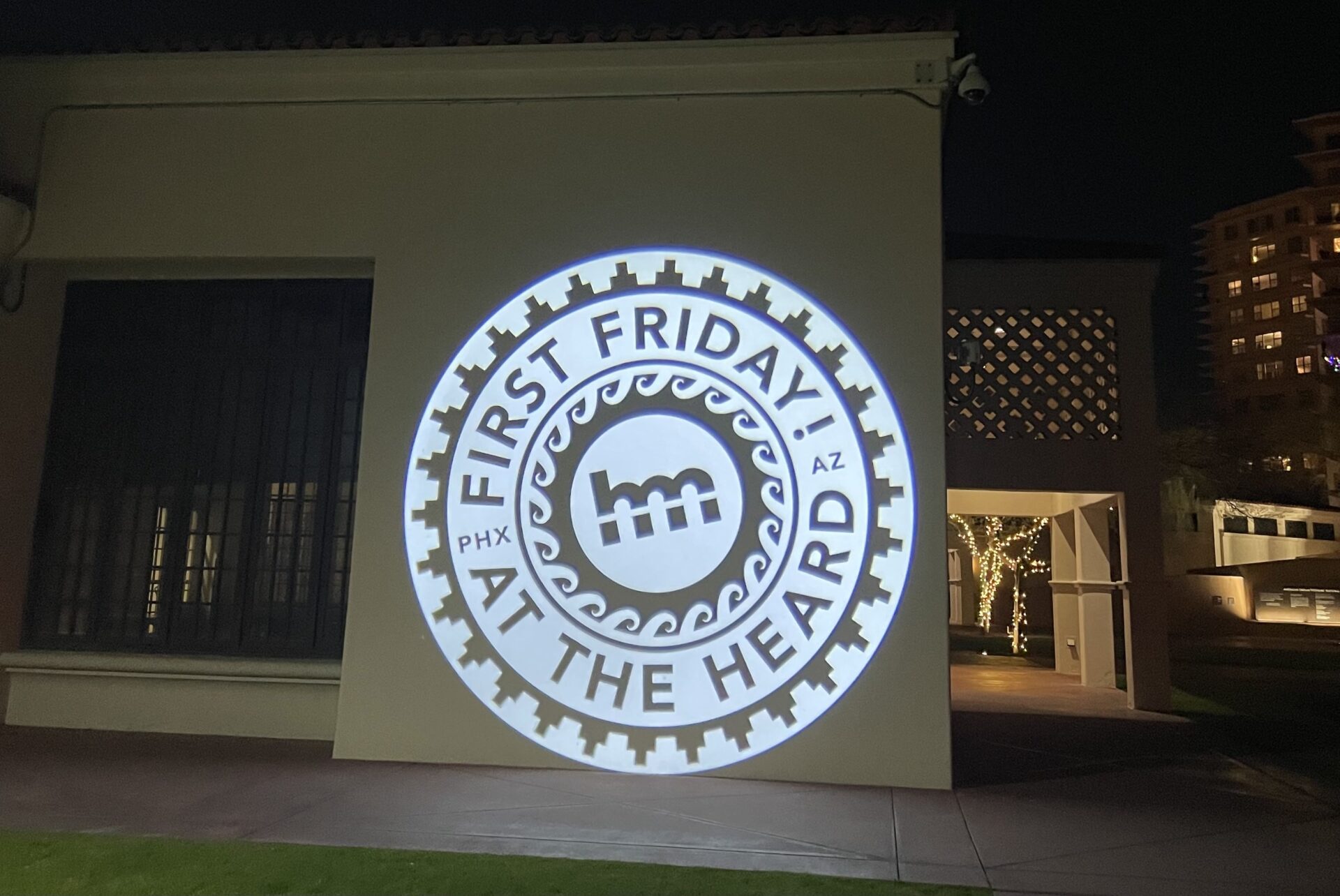 Projection on a building wall showing a circular logo that reads "First Friday! At the Heard, PHX AZ," with a central design element and decorative border.