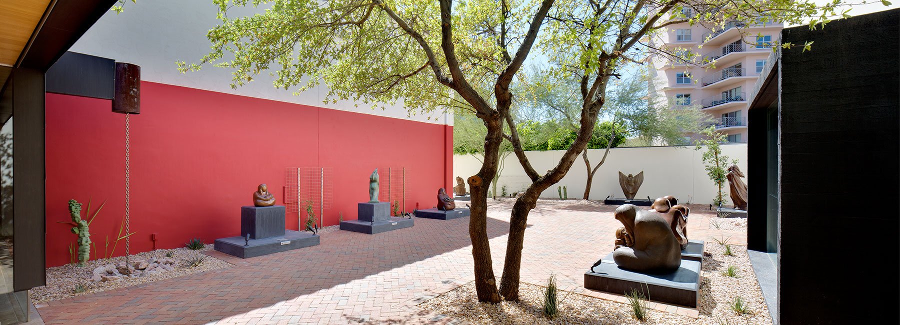 Outdoor sculpture garden with various abstract sculptures on platforms, surrounded by brick pathways, greenery, and a red wall in a sunny setting.