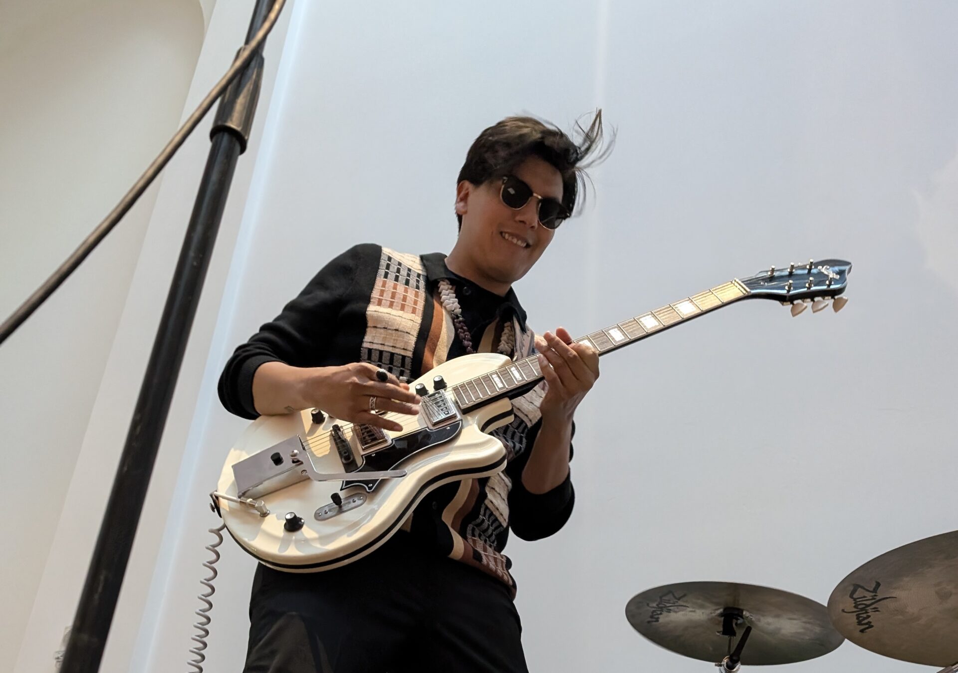 A person in sunglasses plays an electric guitar with a patterned strap. A microphone stand and a cymbal are visible nearby.