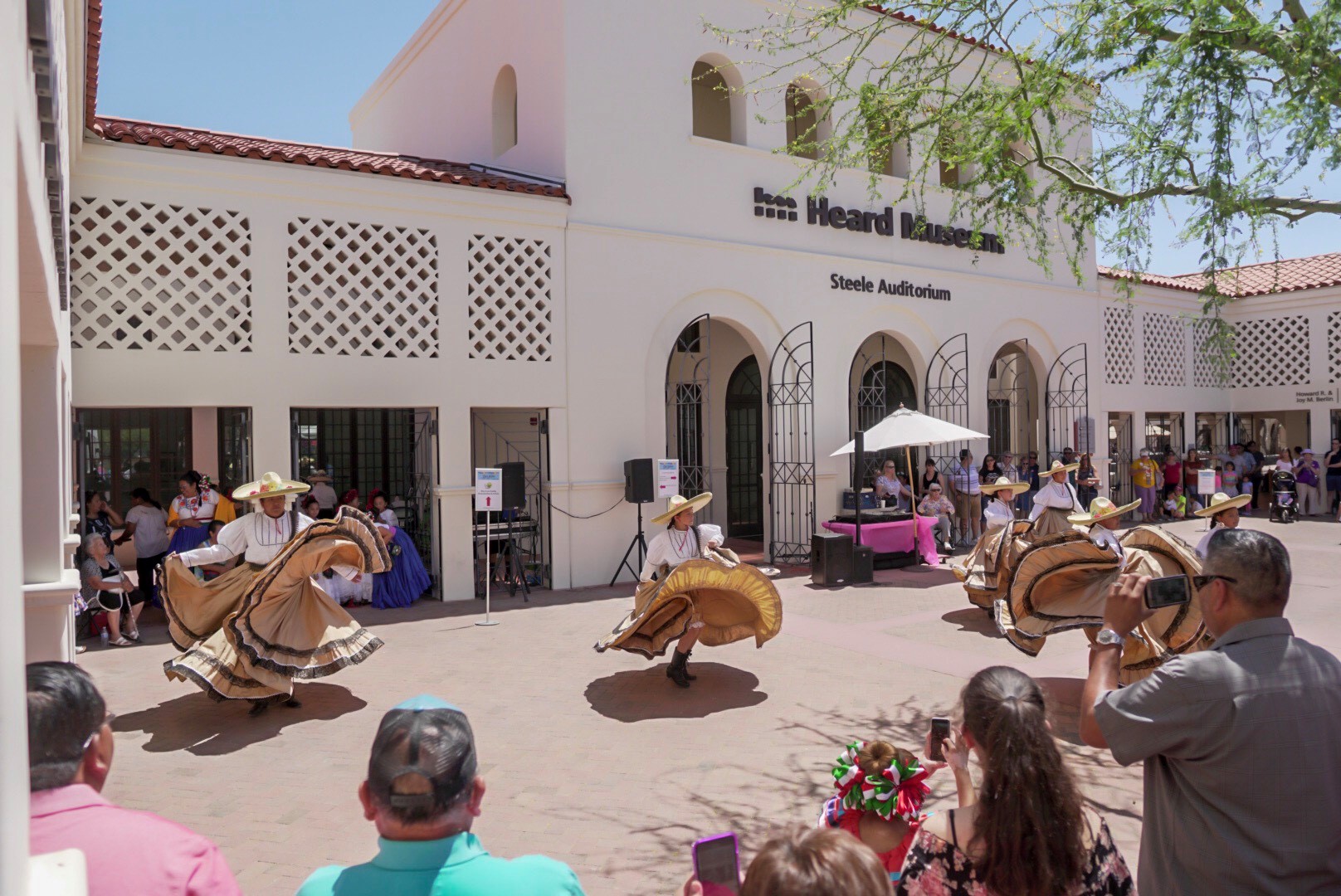 Dancers in traditional attire perform outside the Heard Museum's Steele Auditorium while onlookers watch and take photos.