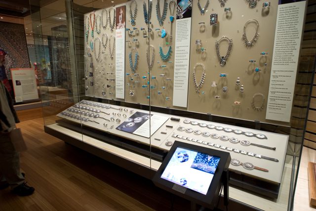 Display case featuring various jewelry pieces including necklaces, bracelets, and rings with turquoise and silver designs. An informational touchscreen is in front of the exhibit.