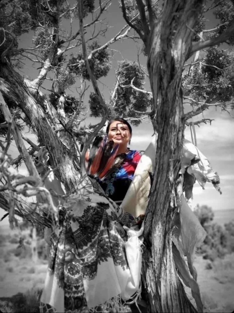 Person in colorful attire stands among the branches of a large tree, holding a fan and looking upward, with sky in the background.