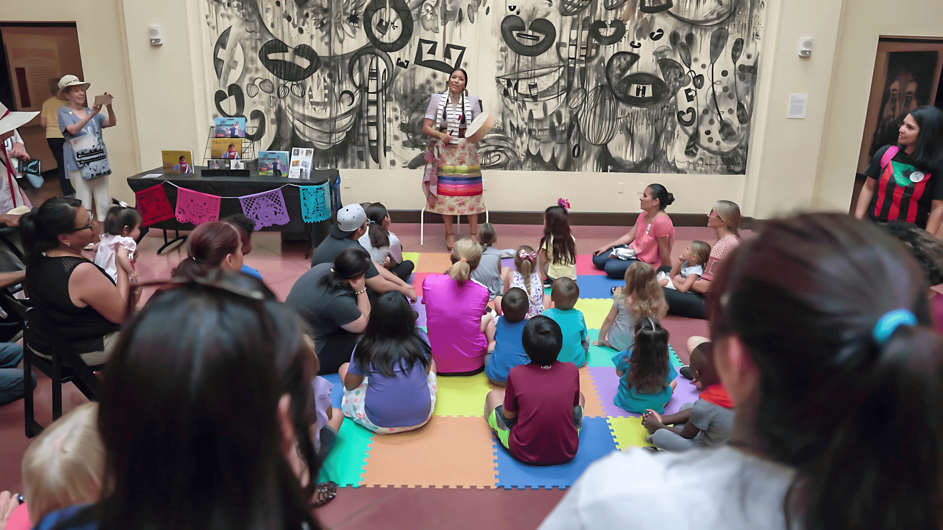 A person stands in colorful attire, speaking to an audience seated on a rainbow mat. A table with crafts is on the side, and a black-and-white mural is on the wall behind.