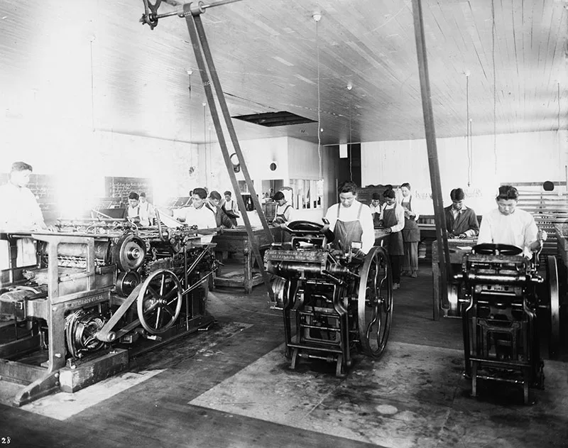 1920 black and white photo of a printing press room with young Native men standing over and working at printing machines.