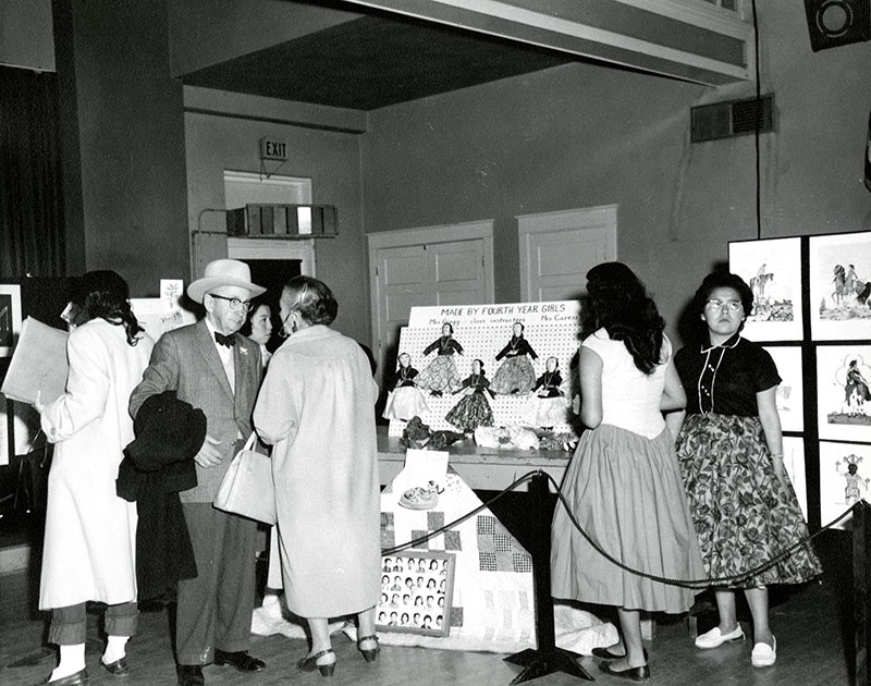 1950 black and white photo of handmade cloth dolls on display at an art event. People mill around looking at the art and talking to each other.