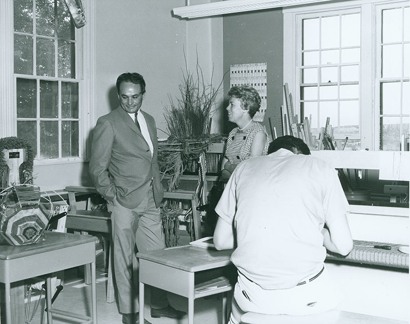 1970 black and white photo of Lloyd Kiva New in a suit standing in weaving classroom with a woman with short blond hair. A young man is hunched over a loom with his back to the camera in the foreground.