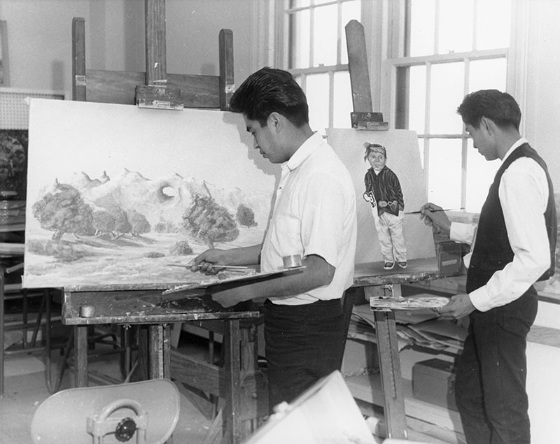 1970 black and white photo of 2 young Native men standing and painting at easels. One paints a landscape and the other paints a portrait of a young boy in traditional Navajo dress.