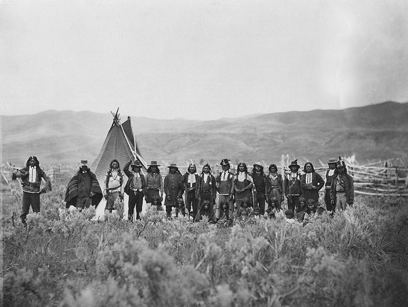 Black and white photo of American Indians in traditional and western dress on the plain in front of a teepee, no date.