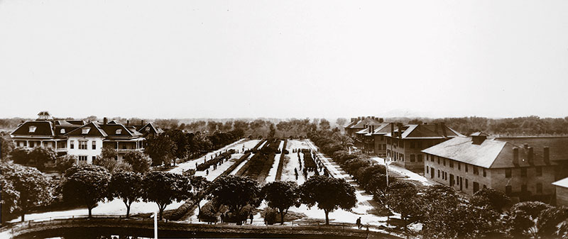 1900 black and white photo aerial view of a center parade grounds with figures lined up and large brick buildings on either side of the grounds.