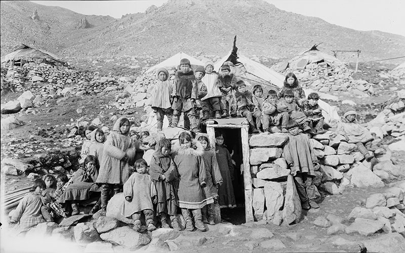 Black and white photo of Alaksan Natives gathered around and on top of a dwelling dug into the ground in a barren rock strewn landscape.