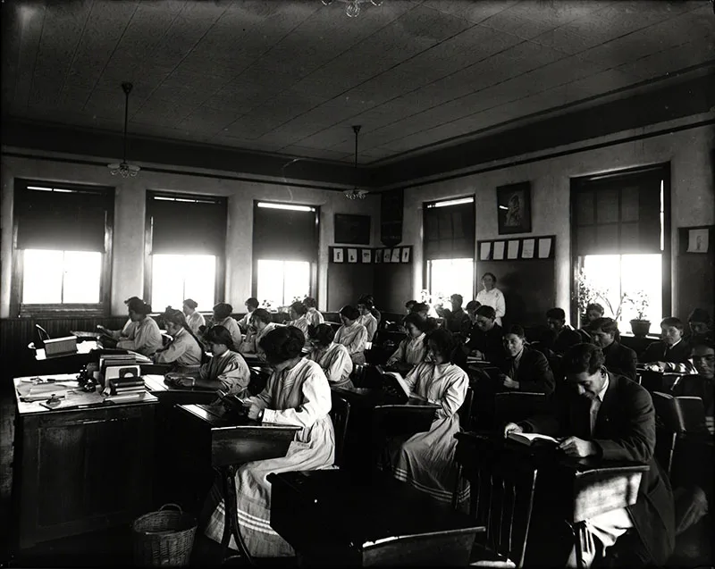 1913 black and white photo of older Native students in a classroom reading at their school desks.
