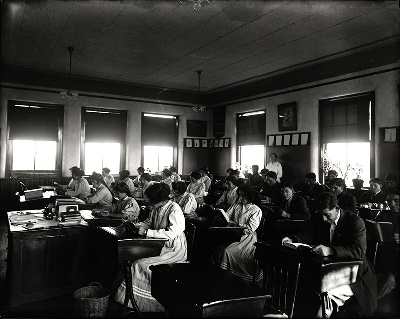 1913 black and white photo of older Native students in a classroom reading at their school desks.