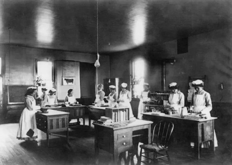 1900 black and white photo of young girls in domestic servant pinafores standing at tables at indistinct activity.