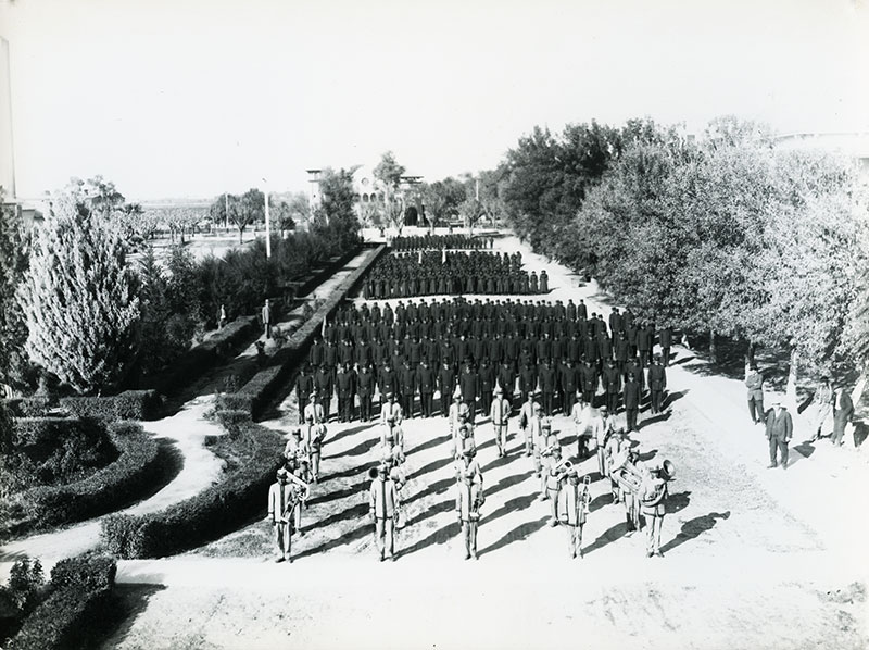 1900 black and white aerial photo of uniformed men and women in formation. A marching band stands in the foreground and the rest of the individuals are in dark uniforms in formation behind them.