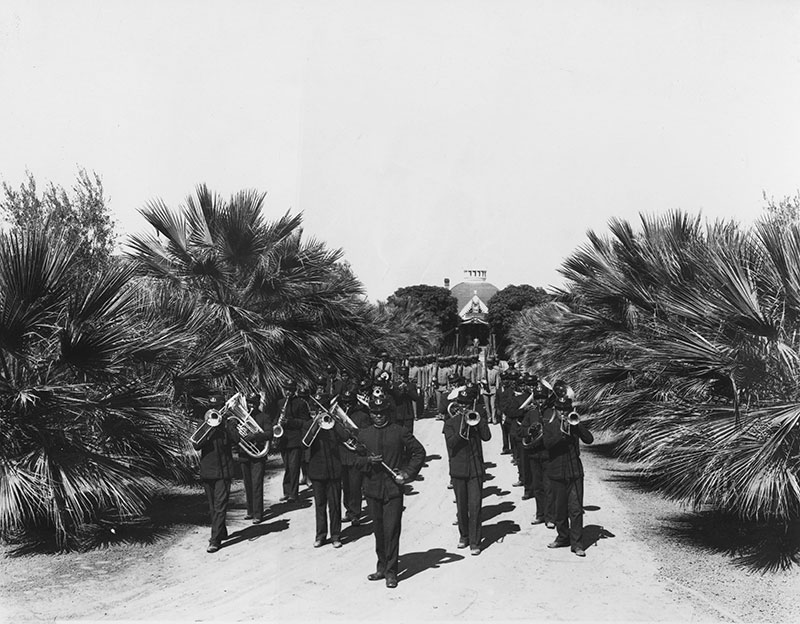 1900 black and white photo of marching band in dark uniforms marching towards the camera on a dirt road between low growing palm trees.