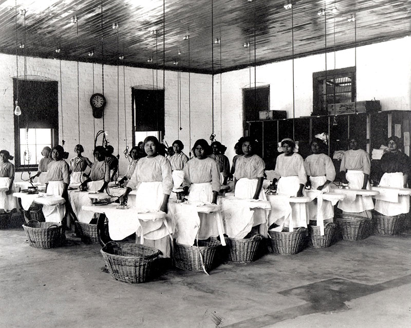 1910 black and white photo of young Native women in a large industrial style laundry room at ironing boards, baskets below.