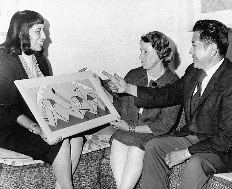 1963 black and white photo of young Native woman happily showing her painting to a Native man and Caucasian woman.