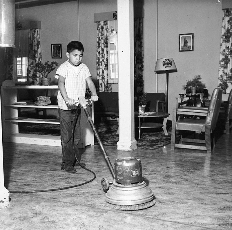 "1950 black and white photo of a young Native boy polishing a floor with a waxing machine in a domestic setting.