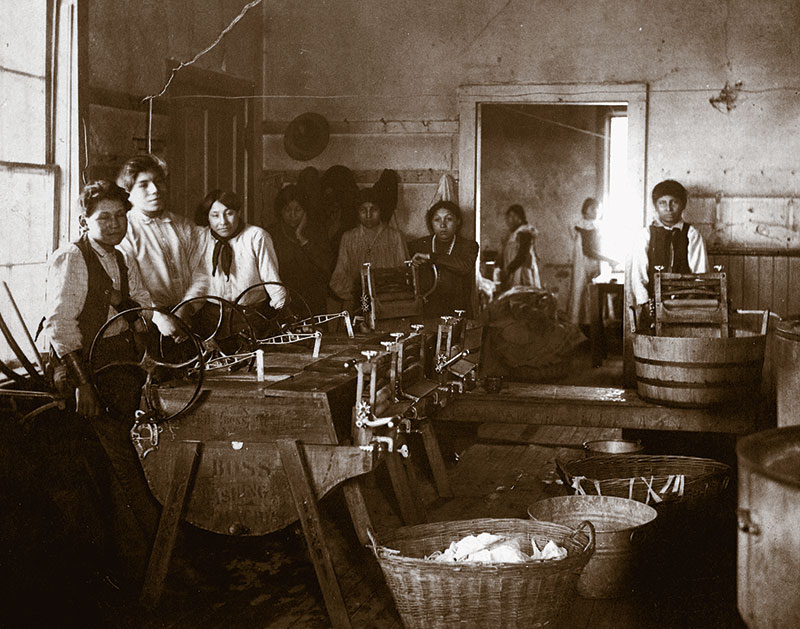 1890 black and white photo of Native youth in laundry room with manual equipment for doing laundry underneath