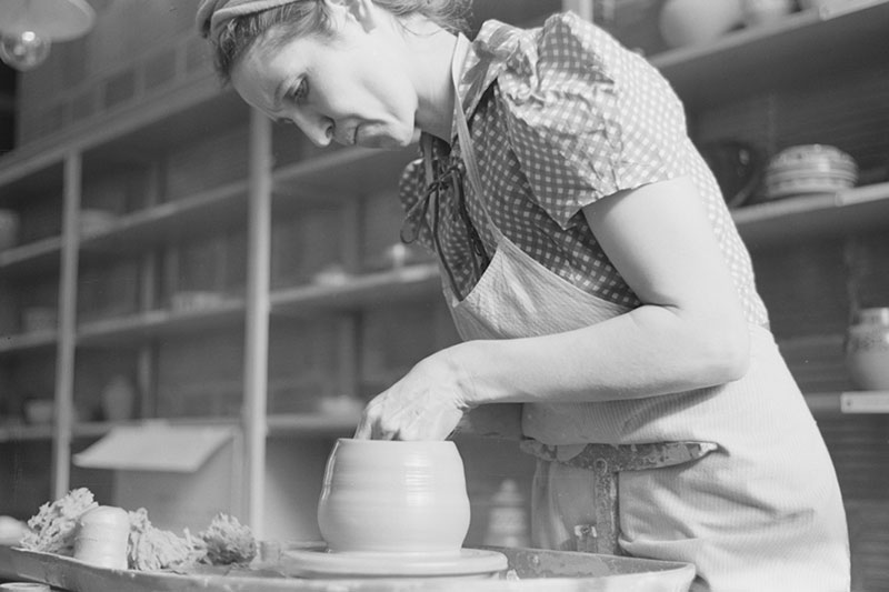 1940s black and white photo of young woman shaping a vessel on a pottery wheel with shelves of pottery behind her.