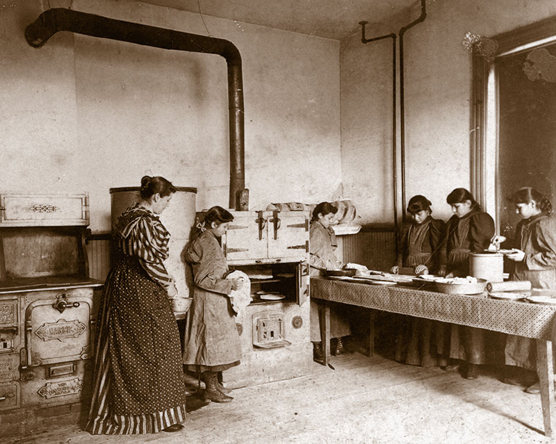 1900 black and white photo of an older woman with several young Native girls baking in a kitchen