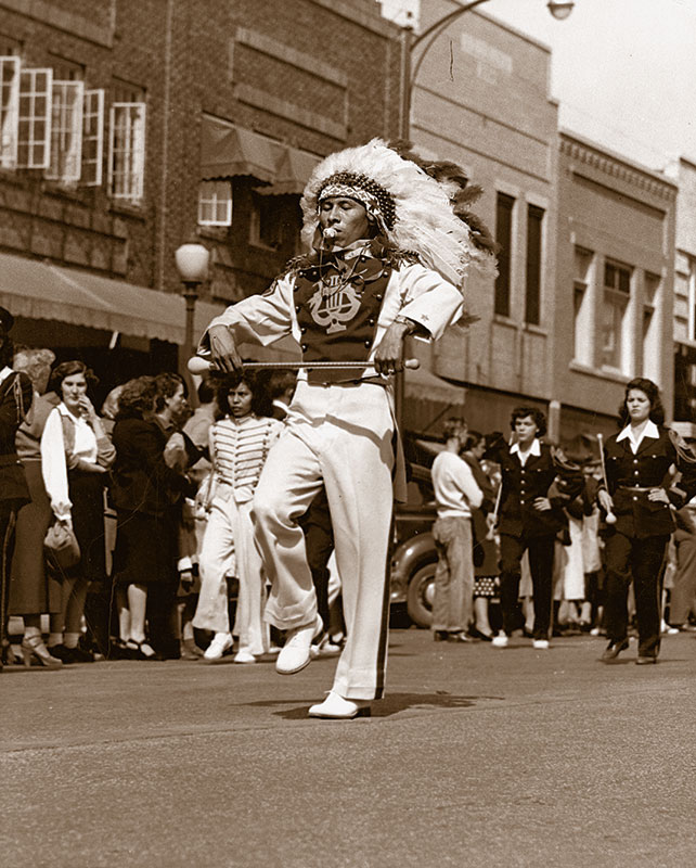 1940 black and white photo of a male drum major in marching costume with headress carrying a twirling batton in both hands and a whistle in his mouth. He marches down the middle of a street in front of the rest of the band with bystanders looking on.